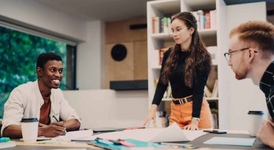 Young cheerful young multiracial students in casual apparel discussing projects at table with cup of coffee documents folders in workplace