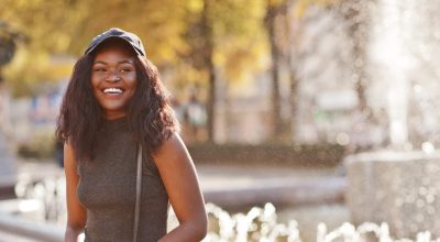 Stylish african american girl in gray tunic, crossbody bag and cap posed at sunny autumn day against fountains. Africa model woman.