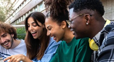 Multiracial college student friends look at mobile phone laughing together. Group of happy people using smartphone outdoors. Youth lifestyle and social media addiction concept.