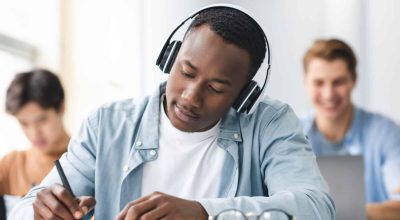 Concetto di studio e apprendimento. Ritratto di giovane uomo nero concentrato con cuffie wireless, seduto al tavolo in un'aula di scuola superiore, che scrive su un quaderno, prendendo appunti o esaminando, con attenzione selettiva.