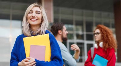 Jeune femme heureuse avec des blocs-notes, souriant et regardant la caméra à l'extérieur de l'université pendant la campagne d'admission.