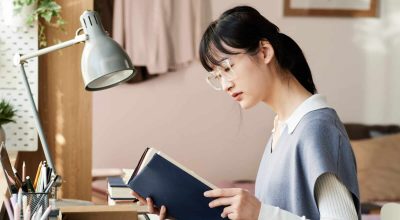 Konzentrierte asiatische Studentin mit Brille und Strickweste sitzt am Tisch und liest ein Lehrbuch, während sie sich auf eine Prüfung vorbereitet