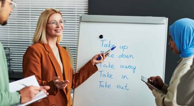 Confident mature teacher of English language pointing at whiteboard with list of phrasal verbs and explaining their meaning to students