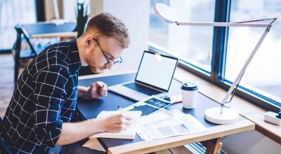 Étudiant caucasien écrivant des notes d'information utiles dans un manuel de connaissances, assis à un bureau de coworking avec un ordinateur portable moderne vierge avec un espace de copie pour un site web international, concept de l'éducation.