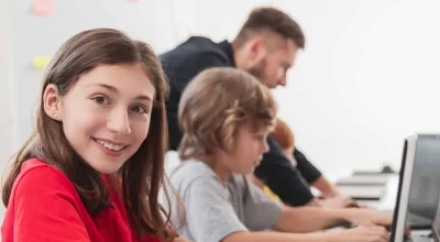 Widok z boku uśmiechniętej nastoletniej uczennicy patrzącej w kamerę podczas lekcji z kolegami z klasy i nauczycielem w nowoczesnej szkole