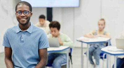 Retrato de um jovem professor afro-americano sentado à secretária e a sorrir para a câmara, com crianças em segundo plano, espaço de cópia