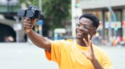 Junger afroamerikanischer Mann, der sich selbst mit einer Videokamera in einer Stadt aufnimmt. Influencer, Reisender, Schöpfer digitaler Inhalte. Foto in hoher Qualität