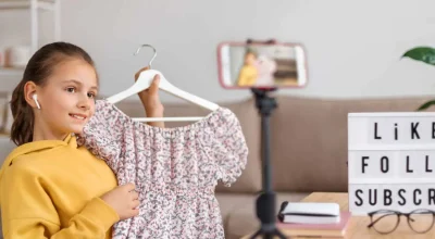 Uma jovem blogueira feliz compartilha sua opinião sobre roupas da moda com seus seguidores, gravando um vídeo com a câmera do smartphone em casa, segurando um vestido, e dá as boas-vindas ao seu canal com um tablet ao fundo